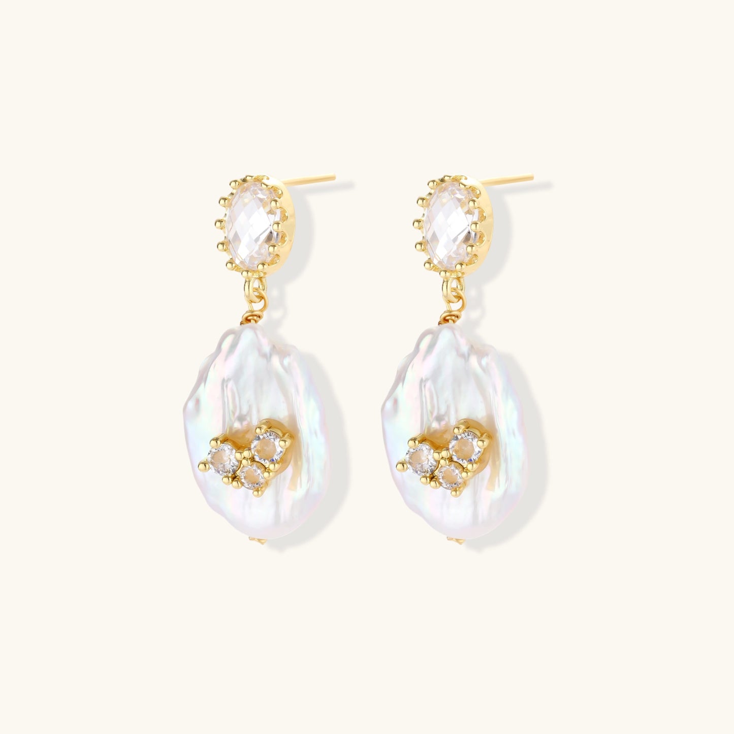 Gold-plated drop earrings with iridescent baroque pearls and crystal flower accents on cream background