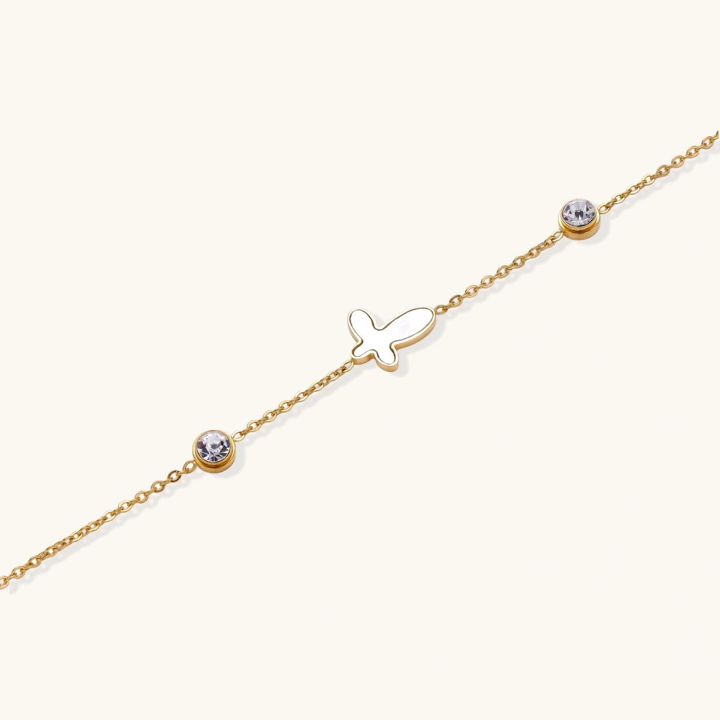 Gold chain bracelet with white mother of pearl butterfly pendant and two bezel-set crystals on cream background